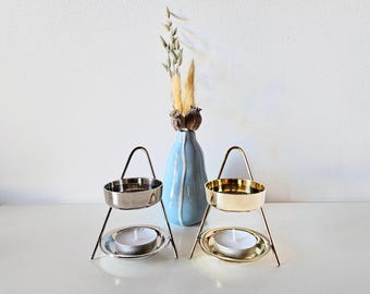 Könnte beinhalten: Zwei Metall-Kerzenhalter, silber- und goldfarben, jeweils mit einer weißen Teelichtkerze. Zwischen ihnen steht eine hellblaue Vase mit getrockneten Blumen. Die Kerzenhalter haben ein minimalistisches, geometrisches Design.