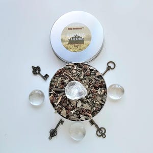 Puede incluir: Una lata plateada abierta, etiquetada "Holy Incenses" y "Elegua Herbs Incense Blend", revela una mezcla de hierbas y un cristal transparente. Llaves decorativas y esferas de cuarzo transparente rodean la lata sobre una superficie blanca.