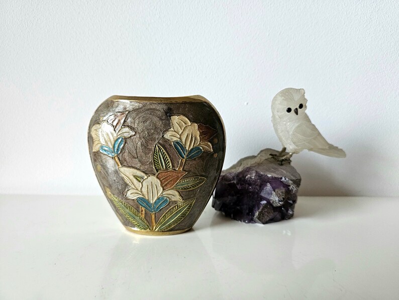 May include: A decorative vase with a floral design in shades of white, blue, and brown. The vase is next to a carved white owl perched on a purple crystal. The vase has a metallic finish.