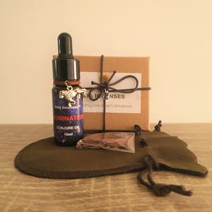 May include: A small glass bottle of "DOMINATION CONJURE OIL" with a black dropper, a silver charm, and a brown leather band. The bottle is next to a small brown box tied with twine, a small bag, and a dark green pouch. The background is a light beige.