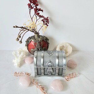 Könnte beinhalten: Eine silberfarbene Metalltruhe mit dekorativen Details, umgeben von rosa Rosenquarzsteinen und Kristallstäben mit Kupferdraht. Eine rote und silberne Vase mit Trockenblumen und eine Geode sind ebenfalls vorhanden.