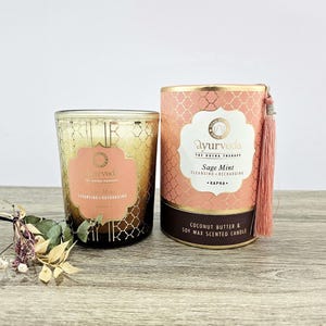 Pode incluir: Duas velas perfumadas, uma em vidro dourado com rótulo rosa, outra em cilindro rosa e dourado com borla rosa. O texto "Ayurveda" e "Sage Mint" é visível. Flores secas em primeiro plano.