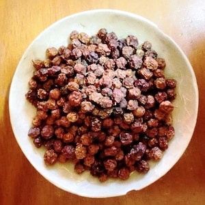 May include: A white ceramic bowl filled with numerous small, round, dried, dark brown berries. The berries have a wrinkled texture and are closely packed. The bowl is on a wooden surface, with the wood grain visible.