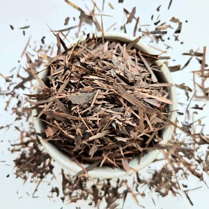 以下が含まれることがあります： 白のセラミックボウルに、茶色の細かく刻まれた樹皮が入っています。樹皮の破片はさまざまなサイズと形状で、白い表面のボウル周辺に散らばっています。画像は真上から撮影されています。