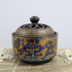Peut inclure: Un brûleur d'encens en céramique avec un couvercle et une base marron foncé, et un corps doré. Le corps présente un motif de dragon détaillé en bleu, rouge et or. Le couvercle a un motif géométrique et un petit bouton. C'est un objet décoratif.