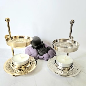 Puede incluir: Dos candelabros de dos niveles, uno dorado y otro plateado, cada uno con una vela de té encendida. Los candelabros tienen un diseño floral en la base y una bandeja perforada en la parte superior. Una pequeña estatua negra de Buda descansa entre ellos.