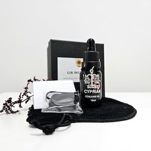 以下が含まれることがあります： 黒いガラス瓶のCyprian Conjure Oil、15ml、スポイト付きが黒いベルベットのポーチの上に置かれています。小さな黒い箱と黒い物体が入った透明なビニール袋がボトルの後ろにあります。ラベルには天秤のデザインがあります。