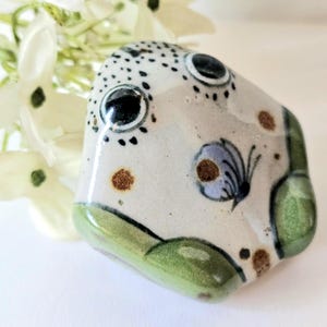 Op de afbeelding: Een keramisch decoratief object met een witte basis, zwarte stippen, bruine vlekken en een blauw vlinderontwerp. Het object heeft een groene basis en staat tegen een achtergrond van witte bloemen.
