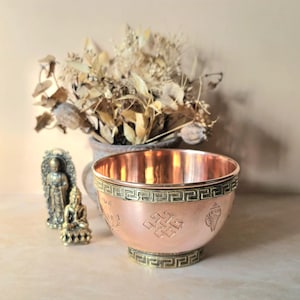 Op de afbeelding: Een koperkleurige kom met een decoratief geometrisch patroon rond de rand en de basis. De kom heeft geëtste symbolen en een gepolijst interieur. Twee kleine Boeddha-beelden en een vaas met gedroogde bloemen staan op de achtergrond.
