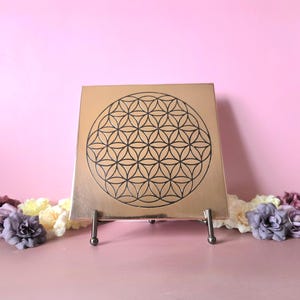 Puede incluir: Placa cuadrada de color cobre con un diseño negro de la Flor de la Vida, exhibida sobre un soporte de metal plateado. La placa está rodeada de flores moradas, amarillas y blancas sobre un fondo rosa.