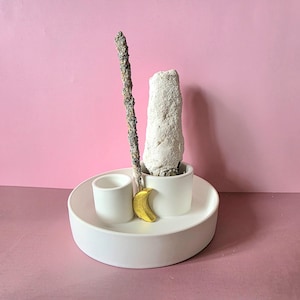 Puede incluir: Un porta incienso de cerámica blanca con base redonda y dos pequeños soportes en forma de taza. Una media luna dorada y dos varillas de incienso se colocan en el soporte. El fondo es de color rosa liso.