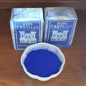 Peut inclure: Deux boîtes rectangulaires étiquetées Azul Castillo Ultramar Puro avec un graphique de château blanc, placées derrière un bol blanc festonné rempli d'un pigment en poudre bleu profond et vibrant. L'emballage est enveloppé dans un film plastique.