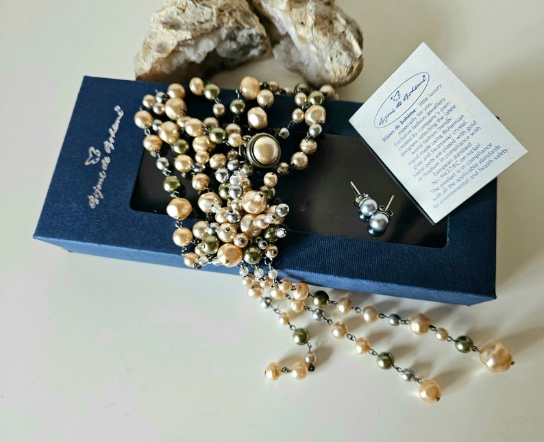 May include: A necklace and earring set with faux pearls in shades of cream, olive green, and silver. The necklace has a large central cluster and cascading strands. The earrings are small, round studs. The set is displayed in a navy blue box.