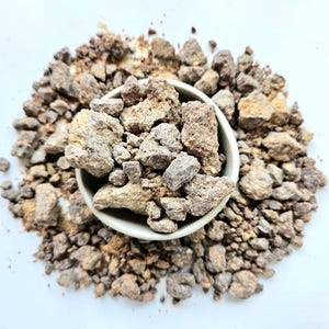 Puede incluir: Vista superior de un pequeño cuenco blanquecino lleno de rocas irregulares de color marrón claro y beige. El cuenco está rodeado por un montón más grande de rocas similares, todo sobre un fondo blanco brillante. Las rocas tienen un aspecto rugoso y texturizado.