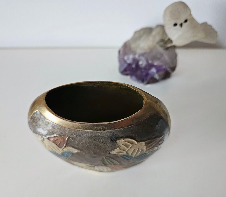 May include: Oval-shaped, dark brown metal bowl with a gold rim. The bowl features a floral design in shades of brown, beige, and blue. A small, white owl figurine sits atop a purple crystal formation in the background.