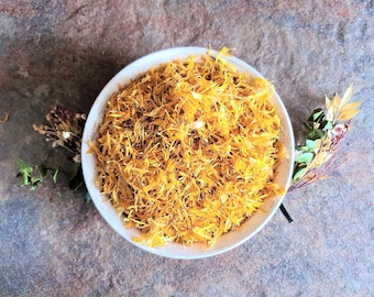 Può includere: Una ciotola bianca piena di petali secchi di calendula arancione. La ciotola è centrata su una superficie screziata grigio-marrone, con rametti di fiori secchi e verde disposti intorno. I petali hanno un aspetto strutturato.