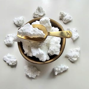 May include: A brass scoop filled with white, irregularly shaped pieces sits in a small, round, bronze-colored bowl. More white pieces are scattered around the bowl on a white surface. The scoop has a leaf design.