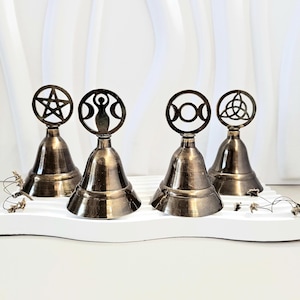 Op de afbeelding: Vier bronskleurige bellen met decoratieve toppen met een pentagram, drievoudige maan, dubbele maan en Keltische knoop. De bellen staan op een wit platform met golvende randen en kleine bloemaccenten.