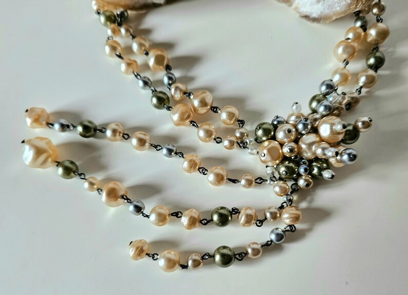May include: A vintage-style necklace featuring multiple strands of faux pearls in cream, silver, and olive green hues. The necklace has a central cluster of pearls and a delicate, cascading design, perfect for adding a touch of elegance to any outfit.