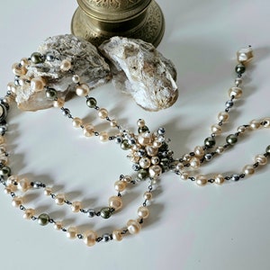 May include: A vintage pearl necklace with a multi-strand design. The necklace features cream, gray, and olive-green pearls strung on a dark chain. A cluster of pearls forms a decorative centerpiece.