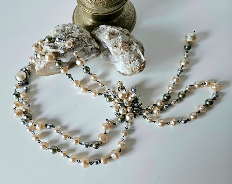 May include: A vintage pearl necklace with a multi-strand design. The necklace features cream, gray, and olive-green pearls strung on a dark chain. A cluster of pearls forms a decorative centerpiece.
