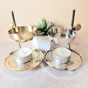 Pode incluir: Dois castiçais de metal, um dourado e outro prateado, cada um com uma pequena vela de chá. Os castiçais têm um design decorativo na base e uma pequena escova presa. Uma pequena planta suculenta está no fundo.