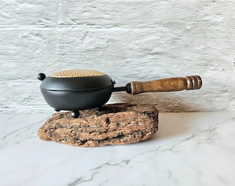 Könnte beinhalten: Kleine, schwarze Gusseisenpfanne mit Holzgriff. Die Pfanne hat eine perforierte Oberseite und steht auf drei kleinen Beinen. Der Griff ist aus hellbraunem Holz. Die Pfanne wird auf einem Holzstück präsentiert.
