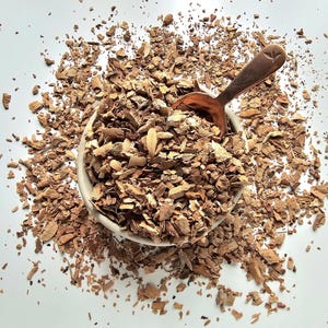 May include: A white bowl filled with brown, shredded bark, with a copper-colored spoon resting inside. The bark pieces are scattered around the bowl on a white surface. The image is taken from a high angle.