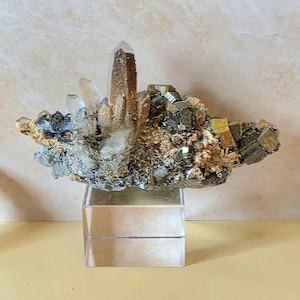 May include: A cluster of crystals, featuring clear quartz points and a central, dark brown crystal. The base is a mix of gold, silver, and tan minerals, displayed on a clear acrylic stand. Decorative crystals are on either side.