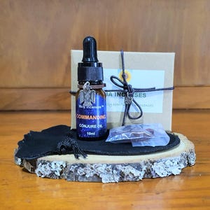 May include: A small glass bottle of "Commanding Conjure Oil" with a black dropper, silver eagle charm, and a blue and purple label. The bottle sits on a wooden slice with a black pouch, a small box, and a clear packet.
