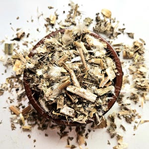 Könnte beinhalten: Eine braune Keramikschale gefüllt mit getrockneten Kräutern und Pflanzenmaterial, einschließlich Stängeln, Blättern und flauschigen Samenkapseln. Die Kräuter sind eine Mischung aus Beige, Braun und Grün, verstreut auf einer weißen Oberfläche.