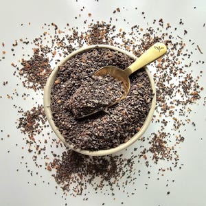 May include: A beige bowl filled with dark brown, granular material, possibly a spice or herb. A gold-coloured spoon rests inside the bowl, partially filled with the same material. Some of the material is scattered around the bowl on a white surface.
