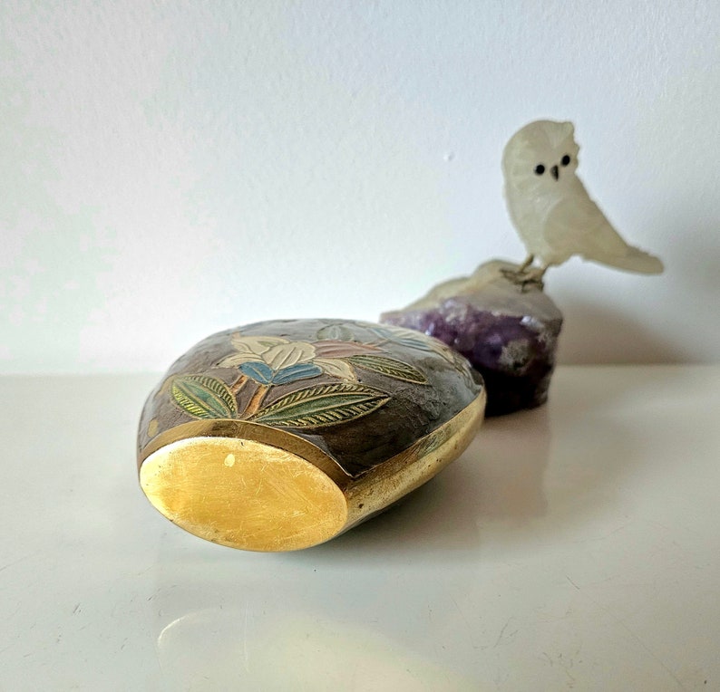 May include: An oval-shaped, dark brown box with floral designs in blue, white, and gold. The box has a gold-colored rim and base. A small, translucent owl figurine sits on a purple rock in the background.
