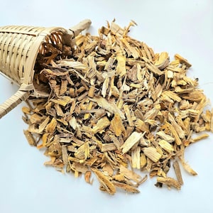 Puede incluir: Una pequeña cesta de bambú trenzado vierte un montón de virutas de madera de color marrón claro sobre una superficie blanca. Las virutas son de varios tamaños y formas, con tonos marrones claros y oscuros. La cesta tiene un asa.