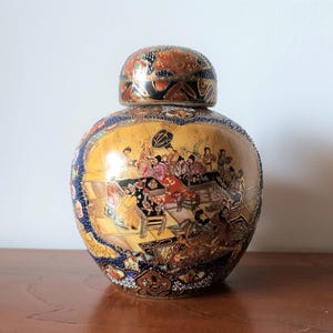 Könnte beinhalten: Eine dekorative Keramikvase mit Deckel, die eine handgemalte Szene von Figuren in einer traditionellen Umgebung zeigt. Die Vase hat einen goldenen Hintergrund mit blauen, roten und schwarzen Akzenten. Der Deckel ist ebenfalls mit komplizierten Mustern verziert.
