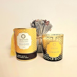 Peut inclure: Deux bougies parfumées et un bouquet de fleurs séchées. L'une est dans un contenant à motifs jaunes avec un pompon, l'autre dans un pot en verre avec un motif doré. Les étiquettes indiquent "Ayurveda Tuberose Jasmine".