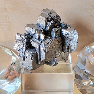 May include: A silver-colored, irregularly shaped sculpture with a textured surface. The sculpture is displayed on a clear, rectangular base, with faceted crystal-like objects on either side. The background is a neutral, light-colored wall.