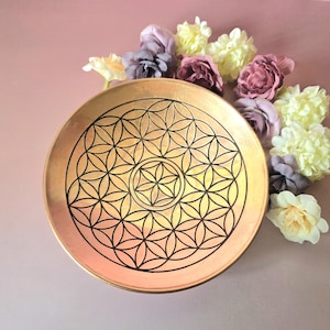 Puede incluir: Un plato decorativo redondo de color dorado con un patrón geométrico negro de la flor de la vida. El plato está rodeado de flores artificiales coloridas en tonos morado, amarillo y rosa, sobre un fondo rosa.