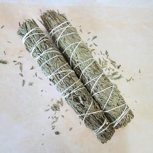 May include: Two bundles of dried sage, bound with white string. The sage is a green-grey colour, with some loose leaves scattered around. The bundles are arranged side by side on a light-coloured surface.