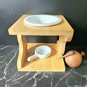 Puede incluir: Una pequeña mesa de madera clara con un cuenco de cerámica blanco en la parte superior y un cuenco más pequeño debajo. Una figura de kiwi de madera descansa a un lado. La mesa tiene un diseño sencillo y elegante.