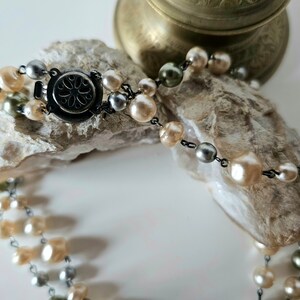 May include: A two-strand pearl necklace with a silver clasp. The pearls are in shades of cream, silver, and olive green. The necklace is displayed on a piece of rough, light-colored stone.