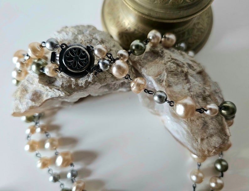 May include: A two-strand pearl necklace with a silver clasp. The pearls are in shades of cream, silver, and olive green. The necklace is displayed on a piece of rough, light-colored stone.
