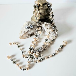 May include: A collection of pearl jewelry, including a necklace and earrings, displayed on a white surface. The necklace features cream, gray, and black pearls. The earrings are small, round, and gray. The jewelry is arranged near a decorative metal object and a piece of stone.