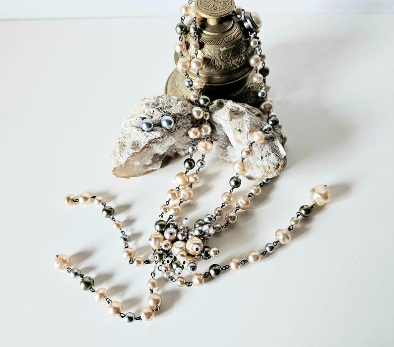 May include: A collection of pearl jewelry, including a necklace and earrings, displayed on a white surface. The necklace features cream, gray, and black pearls. The earrings are small, round, and gray. The jewelry is arranged near a decorative metal object and a piece of stone.
