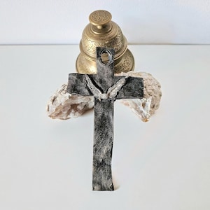 Könnte beinhalten: Ein handgefertigtes, dunkelgraues Kreuz mit strukturierter Oberfläche und detailliertem Taubenemblem. Das Kreuz wird mit einer Messingglocke und einem Stück Naturstein präsentiert, was eine rustikale und spirituelle Ästhetik schafft.