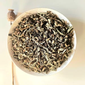 May include: A close-up image of a white ceramic bowl filled with dried herbs. The herbs are a mix of green and brown, with a few stems visible. A dried seed pod is positioned to the left of the bowl.