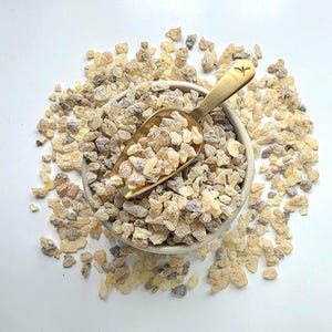 Op de afbeelding: Een kleine, gebroken witte keramische kom gevuld met lichtgeleurde, onregelmatig gevormde harsstukken. Een goudkleurige schep rust in de kom, eveneens gevuld met hars. De kom en de hars zijn omgeven door meer harsstukken op een wit oppervlak.