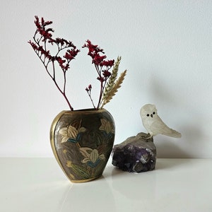 May include: A decorative vase with a floral design and gold trim holds dried red flowers and wheat. A small, white owl figurine sits on a purple amethyst crystal, creating a serene, nature-inspired display.