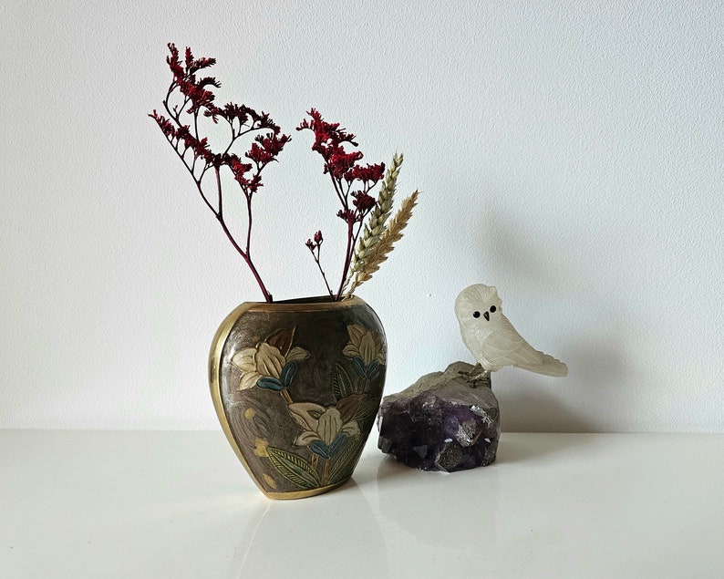 May include: A decorative vase with a floral design and gold trim holds dried red flowers and wheat. A small, white owl figurine sits on a purple amethyst crystal, creating a serene, nature-inspired display.