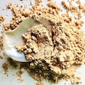 May include: A close-up of a pile of light brown powder with a pearlescent, shell-shaped scoop. The powder is finely ground, likely a cosmetic product such as face powder or bronzer. The background is a plain white surface.